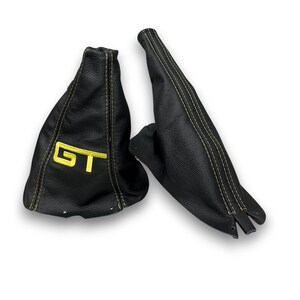 Op de afbeelding: Twee zwarte lederen auto-accessoires met gele stiksels. Eén heeft de gele letters "GT" erop geborduurd. De andere is een lang, smal stuk. Beide lijken nieuw te zijn.