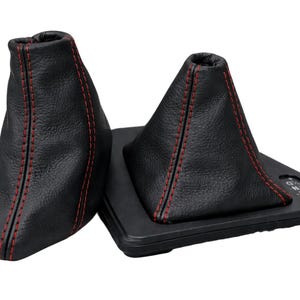 Op de afbeelding: Twee zwarte leren versnellingspookhoezen met rode stiksels. Eén hoes heeft een traditionele vorm, de andere is driehoekig. De hoezen staan op een zwarte basis met versnellingsmarkeringen. Auto onderdelen.