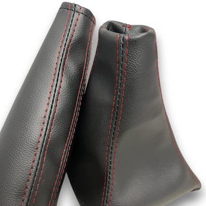 May include: Two black leather car shift boots with red stitching. The boots are made of a smooth, matte leather and have a tapered shape. The red stitching provides a contrasting detail. The background is white.
