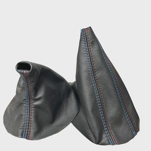 Puede incluir: Dos fundas de palanca de cambios de cuero negro con costuras rojas y azules. Las fundas están hechas de un material de cuero texturizado y están diseñadas para encajar sobre la palanca de cambios de un vehículo.