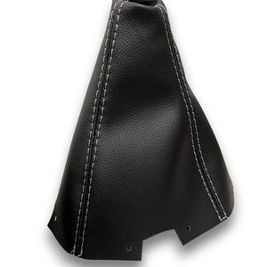 May include: Black leather shift boot with white stitching. The boot has a textured surface and is designed to fit around a vehicle's gear shift lever. The white stitching provides a contrasting detail, and the boot has a trapezoidal shape.