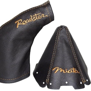 Op de afbeelding: Twee zwarte lederen auto-accessoires, één met "Roadster" geborduurd in goud en de andere met "Miata". Beide hebben gouden stiksels.