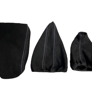 Puede incluir: Tres accesorios interiores de coche de ante negro. Los artículos incluyen una cubierta de consola, una funda de palanca de cambios y una funda de freno de mano. Cada pieza tiene costuras decorativas azules y naranjas.