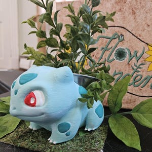 May include: A light blue Bulbasaur planter with green foliage. The planter has teal spots and a red eye. A sign in the background reads "Flower Garden" with a yellow sunflower illustration.