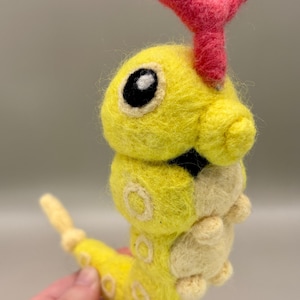 May include: A handmade felt caterpillar toy in yellow, cream, and pink. The caterpillar has a segmented body, large black eyes, and pink antennae. The toy is designed with rounded shapes and soft textures, typical of a felted creation.