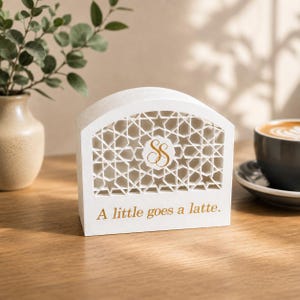 May include: A white arched wooden sign with a laser-cut geometric star pattern and a gold monogram logo in the center. The text "A little goes a latte." is printed in gold lettering at the bottom of the sign, which has a smooth matte finish.
