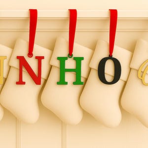 Peut inclure: Cinq bas de Noël crème sont suspendus à une étagère blanche. Chaque bas a un ruban rouge et une lettre : "N" dorée, "N" rouge, "H" verte, "O" noire et "O" dorée. Les lettres sont attachées aux bas avec des rubans rouges ou argentés.