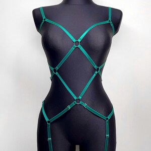 May include: Emerald green body harness with adjustable straps and metal O-rings. The harness is displayed on a black mannequin, showcasing its design and fit. The lingerie piece features a geometric pattern.