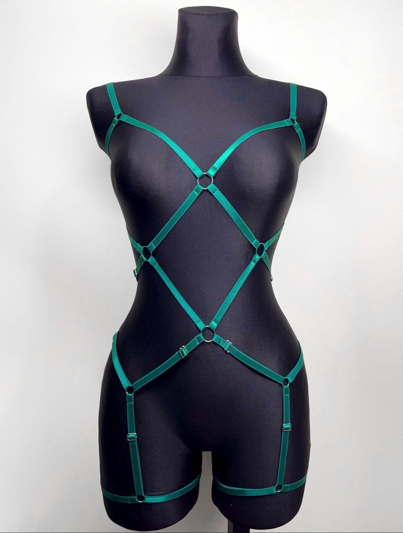 May include: Emerald green body harness with adjustable straps and metal O-rings. The harness is displayed on a black mannequin, showcasing its design and fit. The lingerie piece features a geometric pattern.