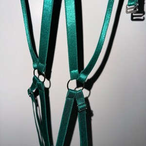 May include: Teal-colored lingerie harness with adjustable straps and metal hardware. The harness features multiple straps connected by metal rings and adjustable buckles. The fabric has a slight sheen, and the design is visible against a white background.