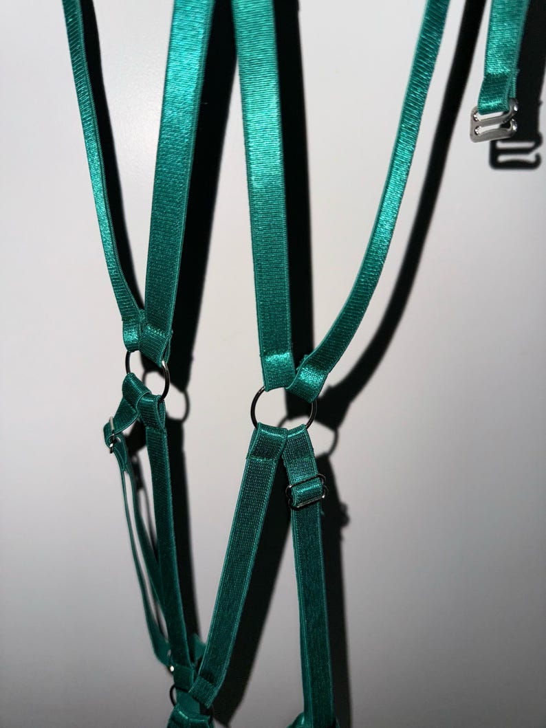 May include: Teal-colored lingerie harness with adjustable straps and metal hardware. The harness features multiple straps connected by metal rings and adjustable buckles. The fabric has a slight sheen, and the design is visible against a white background.