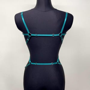 May include: Teal-colored body harness with adjustable straps and metal hardware. The harness features a series of connecting straps that create a geometric design across the torso and back. The garment is displayed on a black mannequin.