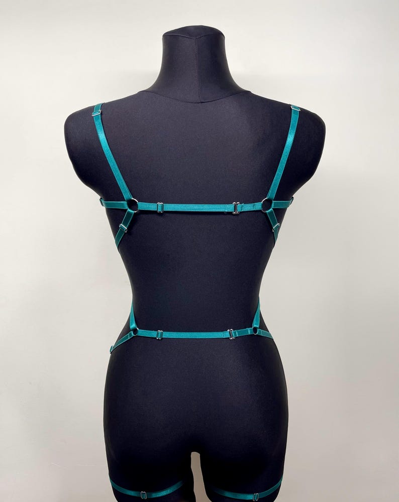 May include: Teal-colored body harness with adjustable straps and metal hardware. The harness features a series of connecting straps that create a geometric design across the torso and back. The garment is displayed on a black mannequin.