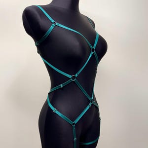 May include: A teal-colored body harness with adjustable straps and metal rings. The harness is displayed on a black mannequin, showcasing its design and fit. The straps create a geometric pattern across the torso and legs.