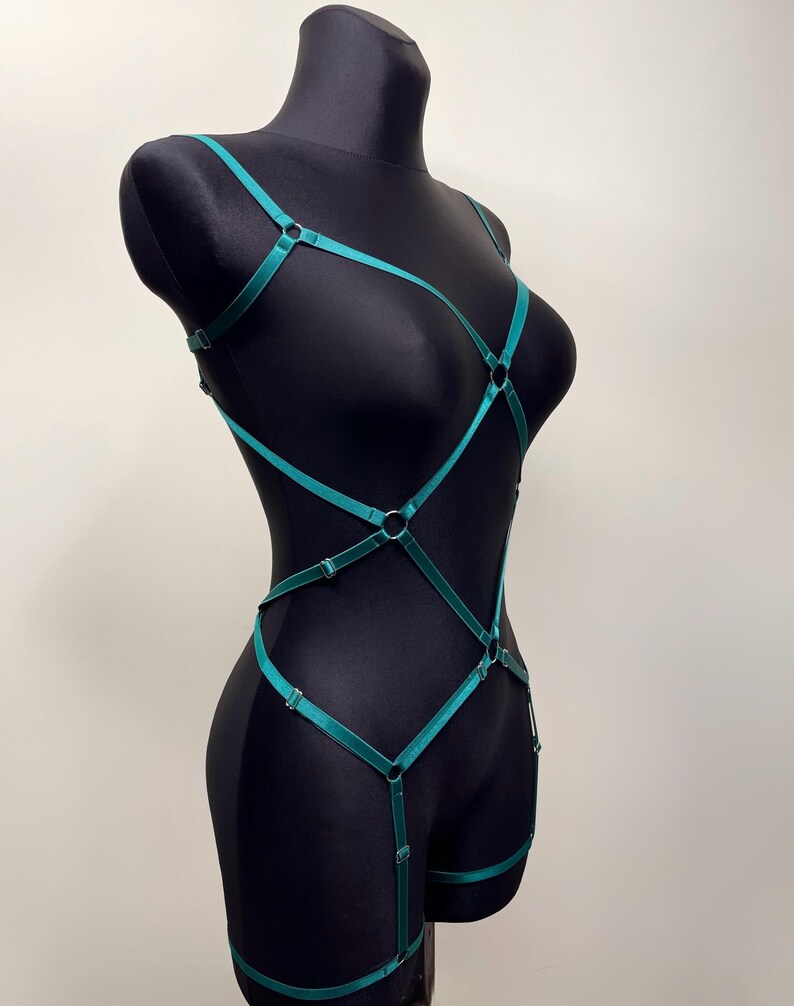 May include: A teal-colored body harness with adjustable straps and metal rings. The harness is displayed on a black mannequin, showcasing its design and fit. The straps create a geometric pattern across the torso and legs.