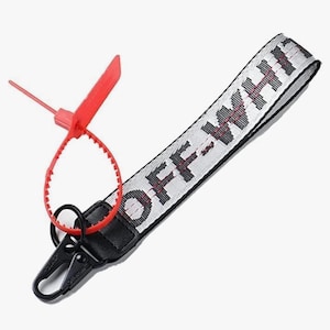 Puede incluir: Correa de muñeca blanca y negra con el texto "OFF-WHITE" en negro. La correa tiene un cierre negro y una etiqueta de plástico roja. Diseñada para llevarse alrededor de la muñeca.