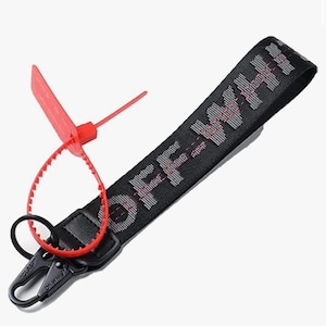 Puede incluir: Correa de muñeca negra y gris con la palabra "OFF WHITE". Cuenta con un cierre negro y una etiqueta de plástico roja. La correa está diseñada para llevarse alrededor de la muñeca, probablemente un accesorio de moda.