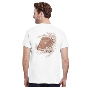 May include: White t-shirt featuring a detailed illustration of a Holy Bible with a cross, surrounded by pink flowers and greenery. The text "Holy Bible" is visible on the cover. A pink ribbon bookmark is also present.