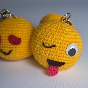 May include: Close-up of two yellow crocheted emoji keychains. One has red heart eyes and a smiling face. The other has a winking eye, a tongue sticking out, and a googly eye. Both have metal key rings.