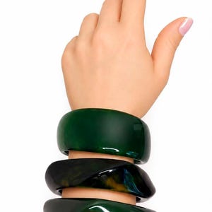 May include: Three stacked bangle bracelets in varying shades of dark green and black. The top bangle is a smooth, solid dark green, while the two lower bangles feature a marbled, translucent resin texture with swirls of deep green, black, and amber.