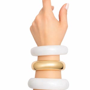 May include: Three bracelets stacked on a wrist. Two are white, and one is gold. The white bracelets are thick and smooth. The gold bracelet is a solid band. The hand is reaching up with the index finger extended.