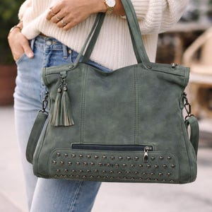 May include: A green suede tote bag with a studded bottom and a zippered front pocket. The bag has a detachable shoulder strap and decorative tassels. The bag is being held by a person wearing a white sweater and blue jeans.