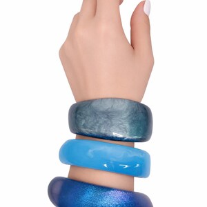 May include: Three chunky bracelets in shades of blue adorn a wrist. The top bracelet is a marbled teal, the middle is a solid light blue, and the bottom is a glittery dark blue. Jewelry.