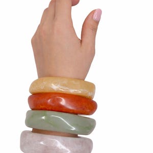 May include: A stack of four colorful jade bracelets in shades of yellow, orange, green, and pink. The bracelets are smooth and rounded, stacked on a wrist against a white background.
