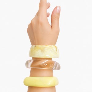 May include: A set of three chunky bangle bracelets worn on the wrist. The top bangle is a marbled cream and yellow resin, the middle is a clear translucent brown resin, and the bottom bangle is a solid opaque pale yellow resin with a smooth finish.