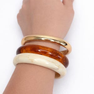 May include: A collection of three bangles in varying colors. The top bangle is gold, the middle one is a marbled brown, and the bottom one is off-white. The bangles are stacked on a wrist against a white background.