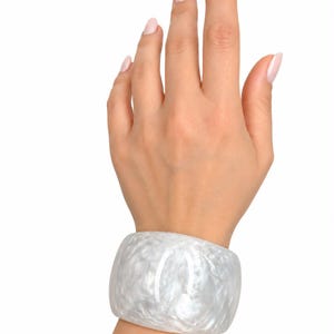 May include: A wide, translucent white bangle bracelet with a marbled, pearlescent texture. The bracelet has a smooth, polished finish and a thick, rounded band design that encircles the wrist, showcasing a cloudy, swirling pattern throughout.