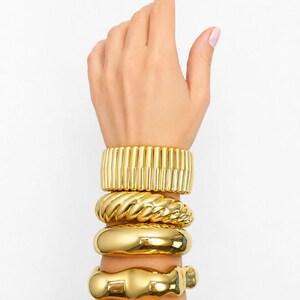 May include: A stack of four gold-toned metal bracelets worn on the wrist. The set includes a ribbed cuff, a twisted rope-textured band, a smooth polished thick bangle, and a final bangle with an irregular, organic, and wavy molten-like shape.