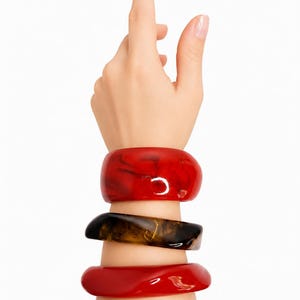 May include: A set of three chunky bangle bracelets worn on the wrist. The top bracelet is a glossy, translucent red with darker swirls. The middle bracelet features a marbled brown and black pattern. The bottom bracelet is a solid, glossy red.