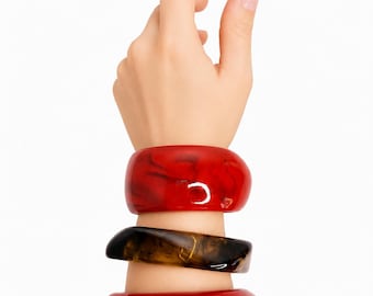 May include: A set of three chunky bangle bracelets worn on the wrist. The top bracelet is a glossy, translucent red with darker swirls. The middle bracelet features a marbled brown and black pattern. The bottom bracelet is a solid, glossy red.