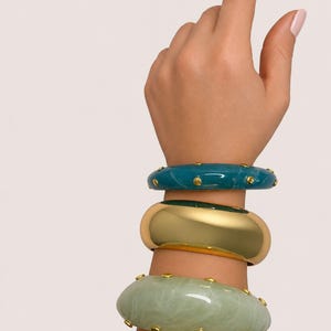 May include: Three bracelets stacked on a wrist. The top bracelet is teal with gold studs. The middle bracelet is gold. The bottom bracelet is light green with gold studs. The bracelets are a mix of solid and marbled designs.
