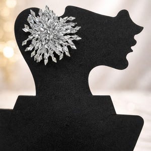 May include: A sparkling silver-colored brooch in a starburst design. The brooch is composed of many small, clear crystals. It is displayed on a black mannequin head, showcasing its intricate details and elegant design. The background is blurred.