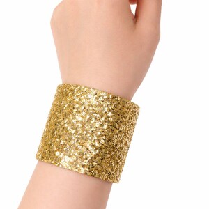 May include: A wide, gold glitter cuff bracelet. The cuff is wrapped around a wrist and is made of small, sparkling gold sequins. The bracelet is a statement piece, perfect for adding a touch of glamour to any outfit.