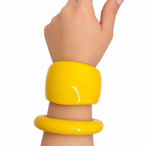 May include: Two bright yellow bangles are worn on a wrist. The top bangle is a wide, rectangular shape, while the bottom bangle is a rounded, circular shape. The bangles have a glossy finish and reflect light. The background is plain white.