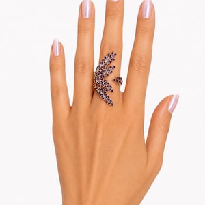 May include: A gold-toned ring featuring a large, leaf-shaped design adorned with numerous marquise-cut purple gemstones. The ring spans across the middle finger, with a smaller, matching leaf-shaped accent piece positioned on the ring finger.