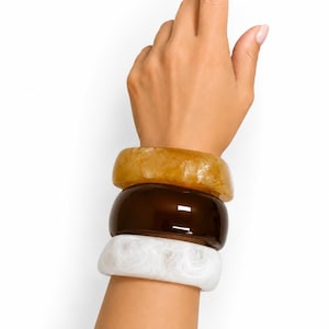 May include: A stack of three chunky bracelets in shades of amber, brown, and white. The amber bracelet is at the top, followed by a dark brown one, and a white marbled design at the bottom. The bracelets are worn on a wrist, with a hand pointing upwards.