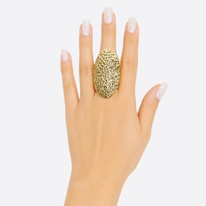 May include: A large, oval-shaped gold ring featuring a textured, hammered surface. The ring has a prominent, elongated design that covers a significant portion of the finger, showcasing a reflective metallic finish with irregular indentations.