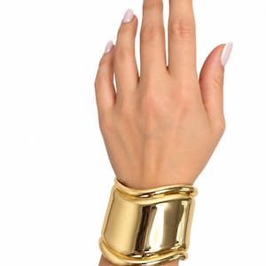 May include: A wide, polished gold-tone cuff bracelet featuring a smooth, reflective surface with undulating, wave-like edges at the top and bottom. The metal has a high-shine finish that catches the light, creating bright highlights across the curved form.