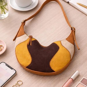 May include: A patchwork shoulder bag in brown, tan, cream, and yellow. The bag has a curved shape with a brown leather strap and a small zipper pull. The bag is on a light-colored wooden surface, with a cup of coffee, a notebook, and makeup nearby.