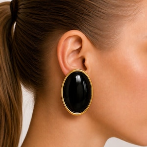 May include: Close-up of a large, oval-shaped black earring with a gold-colored border. The earring is worn on the ear, showcasing its glossy, polished surface. The design is simple yet elegant, suitable for various occasions.