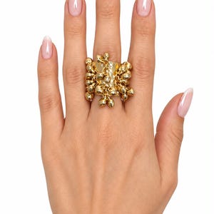 May include: A wide gold-tone ring featuring a textured band adorned with numerous small, dangling metallic beads that create a layered, fringe-like effect across the surface of the jewelry piece, reflecting light with a polished, reflective finish.