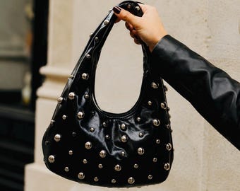 May include: Black handbag with silver-colored studs. The bag has a curved shape and a short handle. The bag is held up by a person wearing a black leather jacket.