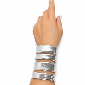 May include: A set of four silver-toned cuff bracelets with a smooth, reflective, and irregular wavy surface. Each bracelet features a wide, flat band design that wraps around the wrist, creating a layered appearance with polished metallic edges.
