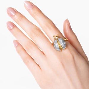 May include: A gold-toned ring in the shape of a ladybug, encrusted with sparkling white and yellow gemstones. The ring's design features a split body, with the wings open. The ring is worn on a finger.