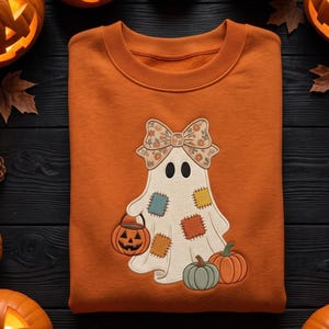 May include: An orange crewneck sweatshirt featuring an embroidered white ghost character. The ghost wears a floral patterned bow and has colorful square patches. It holds a small jack-o'-lantern bucket and stands beside two pumpkins.