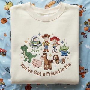 以下が含まれることがあります： クリーム色のスウェットシャツ。刺繍デザインは、映画「トイ・ストーリー」のキャラクター。ウッディ、バズ・ライトイヤー、その他のキャラクターが含まれ、下に「You've Got a Friend in Me」のフレーズがあります。